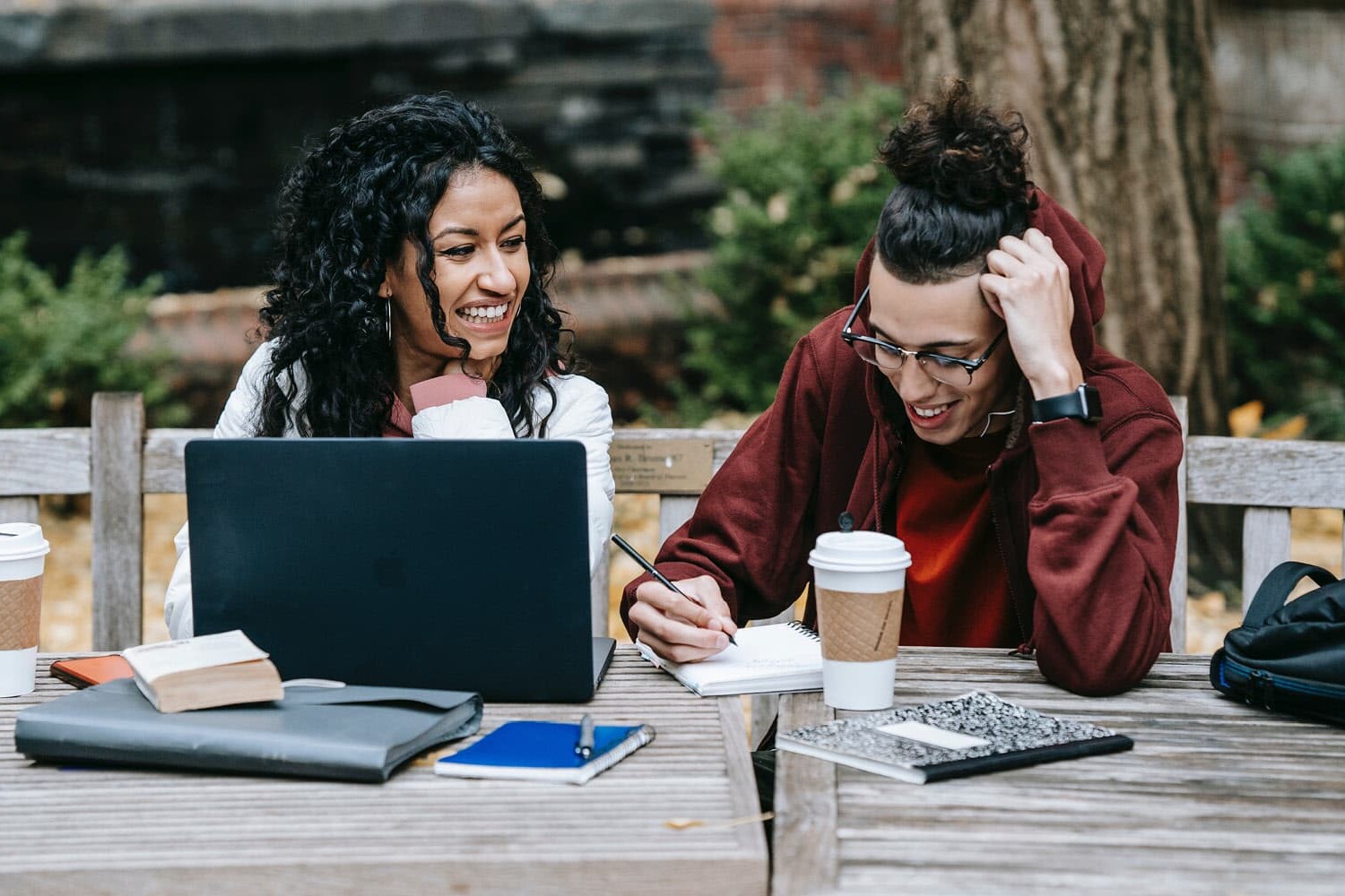 Two people sit at an outdoor wooden table with a laptop, notebooks, and coffee cups, smiling and working together. Surrounded by trees and greenery, their collaboration reflects how youth see hope for reconciliation—but know it will take work.