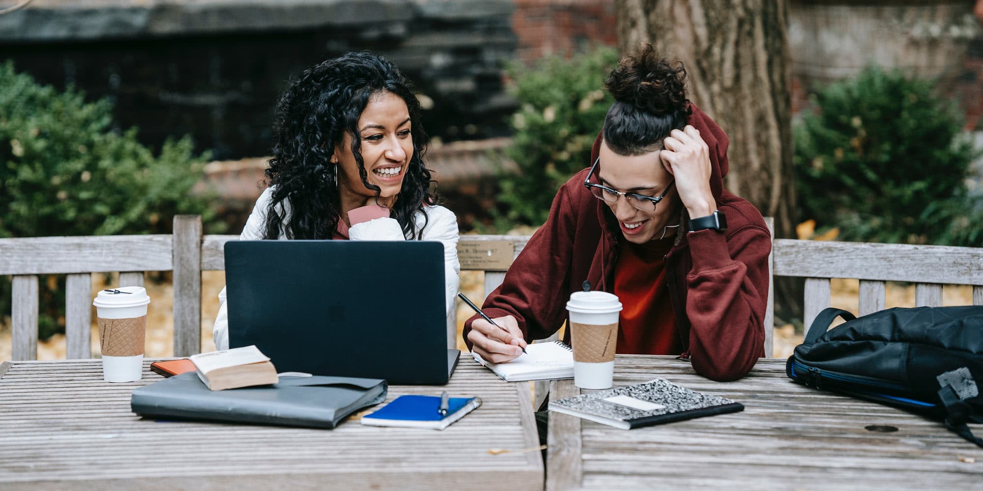 Two people sit at an outdoor wooden table with a laptop, notebooks, and coffee cups, smiling and working together. Surrounded by trees and greenery, their collaboration reflects how youth see hope for reconciliation—but know it will take work.