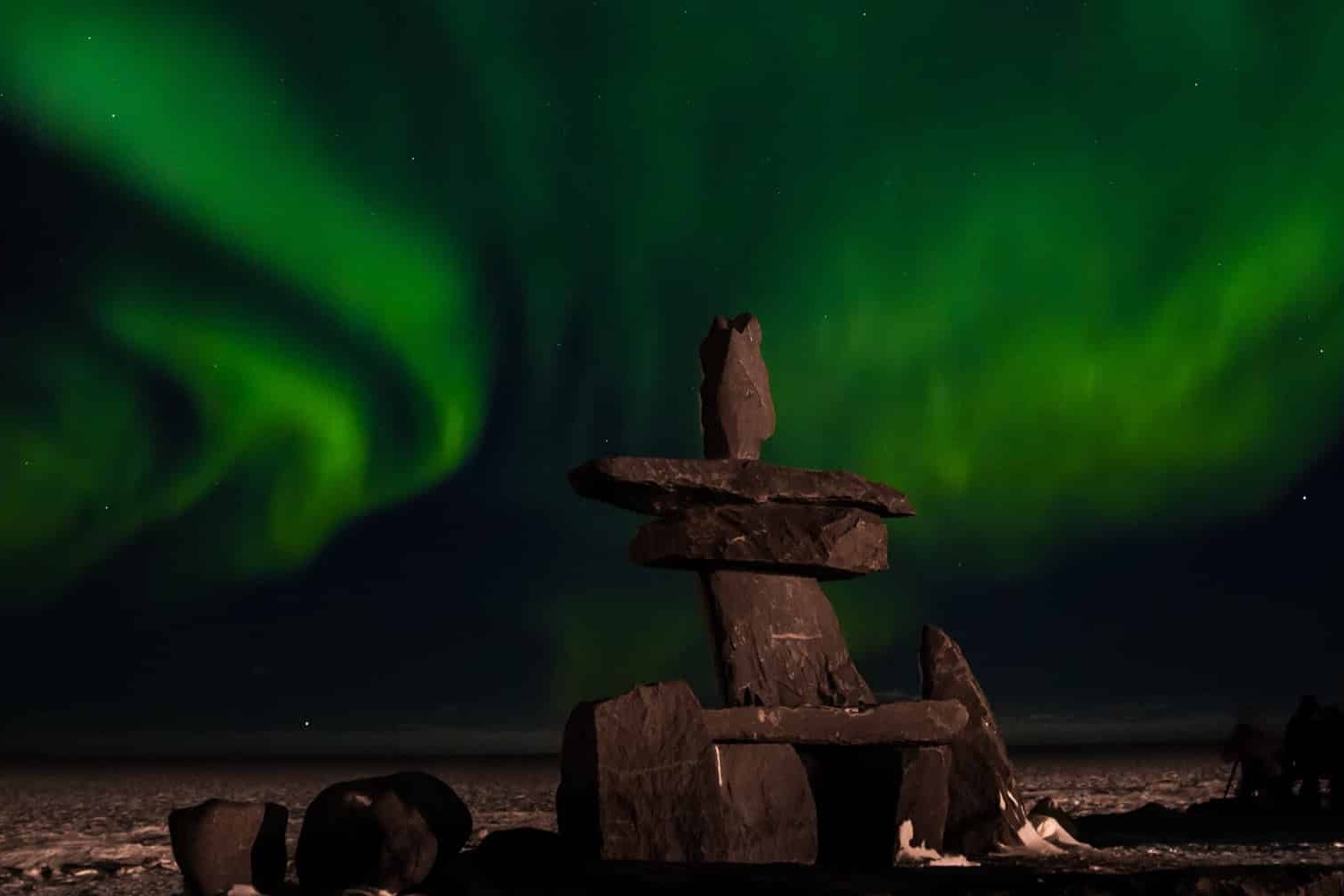 A large stone inukshuk stands on snowy ground at night, with vibrant green northern lights swirling in the dark sky above, symbolizing that youth see hope for reconciliation but it will take work.