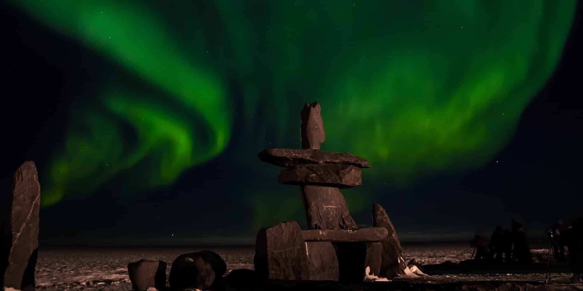 A large stone inukshuk stands on snowy ground at night, with vibrant green northern lights swirling in the dark sky above, symbolizing that youth see hope for reconciliation but it will take work.