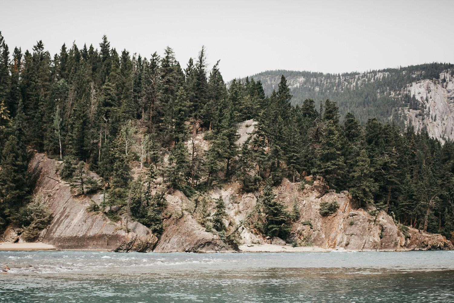 A turquoise river flows past rocky cliffs covered with dense pine trees, with a forested mountain rising in the background under a cloudy sky—reflecting how coronavirus will not dent the trust Canadians have in each other.