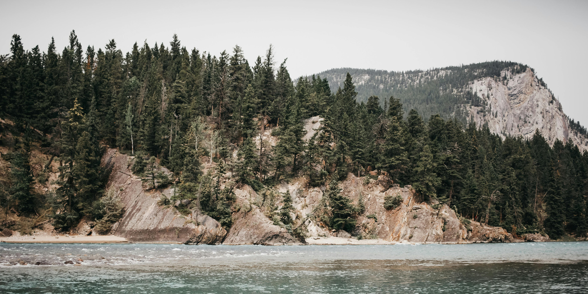 A turquoise river flows past rocky cliffs covered with dense pine trees, with a forested mountain rising in the background under a cloudy sky—reflecting how coronavirus will not dent the trust Canadians have in each other.