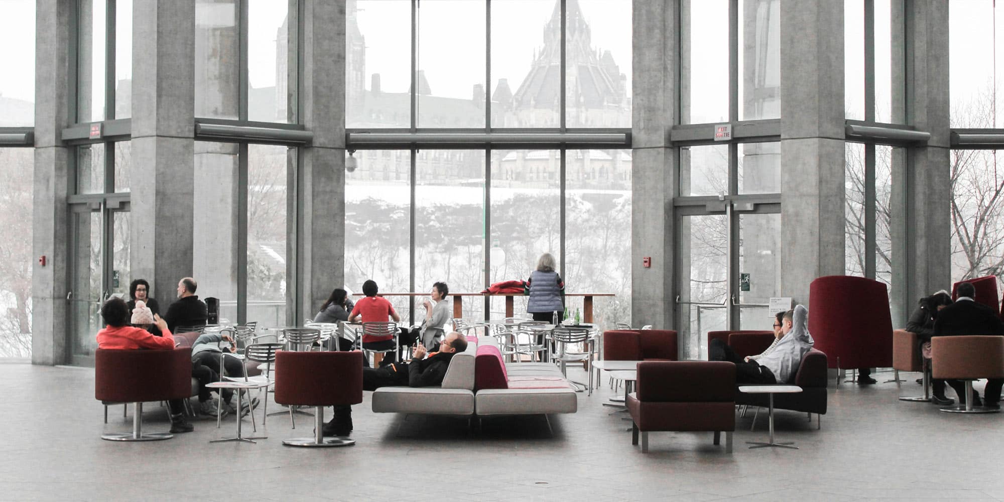 People sit and talk in a spacious, modern lounge with large windows overlooking a snowy landscape and a historic building. Despite recent challenges, Coronavirus will not dent the trust Canadians have in each other as they relax or work together.