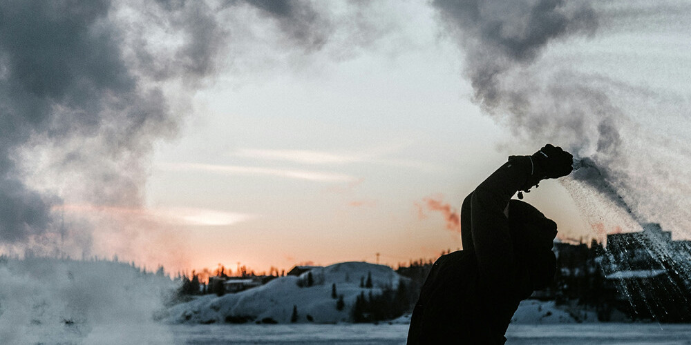 A person throws hot water into cold air outdoors, creating a dramatic cloud of steam that arches above them against a snowy landscape at sunset—a scene reminiscent of an Environics Institute Work exploring natural phenomena.