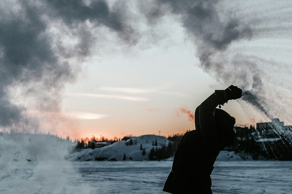 A person throws hot water into cold air outdoors, creating a dramatic cloud of steam that arches above them against a snowy landscape at sunset—a scene reminiscent of an Environics Institute Work exploring natural phenomena.
