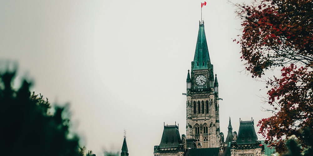 The Parliament building in Ottawa, Canada, with its clock tower and Canadian flag on top, is seen through trees with autumn leaves under an overcast sky—a scene often featured in Environics Institute Work exploring Canadian identity.