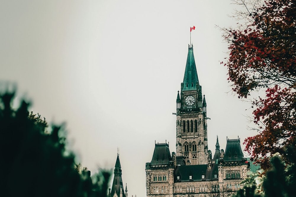 The Parliament building in Ottawa, Canada, with its clock tower and Canadian flag on top, is seen through trees with autumn leaves under an overcast sky—a scene often featured in Environics Institute Work exploring Canadian identity.