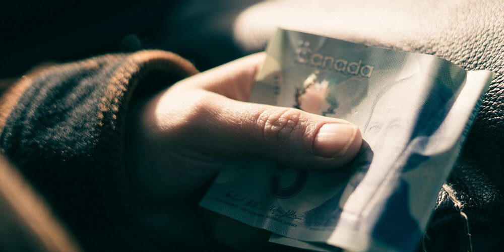 A hand holding a crumpled Canadian five-dollar bill over a dark leather bag, partially visible in soft, natural light—an everyday moment reminiscent of Environics Institute Work themes and insights.