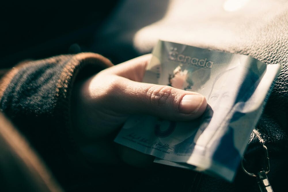 A hand holding a crumpled Canadian five-dollar bill over a dark leather bag, partially visible in soft, natural light—an everyday moment reminiscent of Environics Institute Work themes and insights.