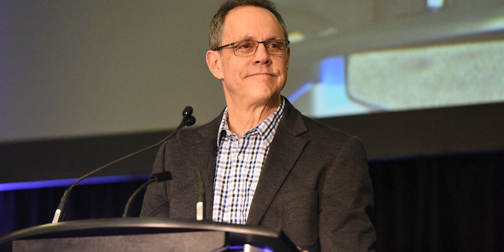 A middle-aged man wearing glasses, a checked shirt, and a dark blazer stands at a podium with microphones, smiling slightly. A large screen in the background hints at the question: Is Canada as a beacon of migrant acceptance still deserved?.