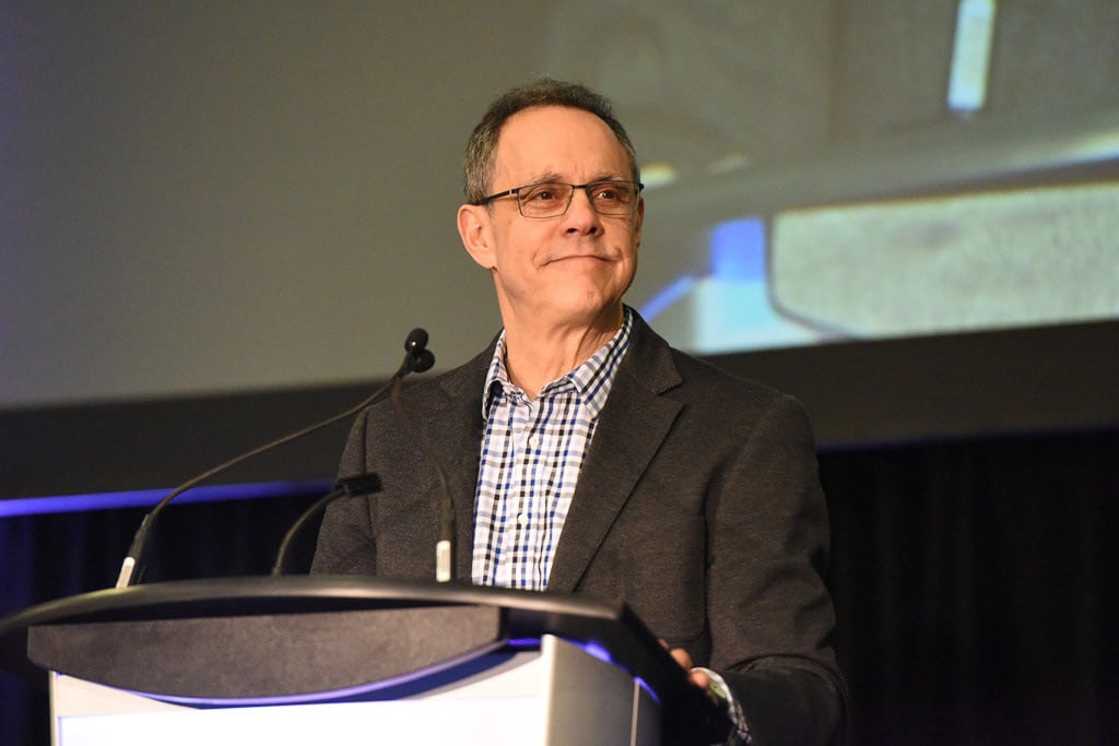 A middle-aged man wearing glasses, a checked shirt, and a dark blazer stands at a podium with microphones, smiling slightly. A large screen in the background hints at the question: Is Canada as a beacon of migrant acceptance still deserved?.