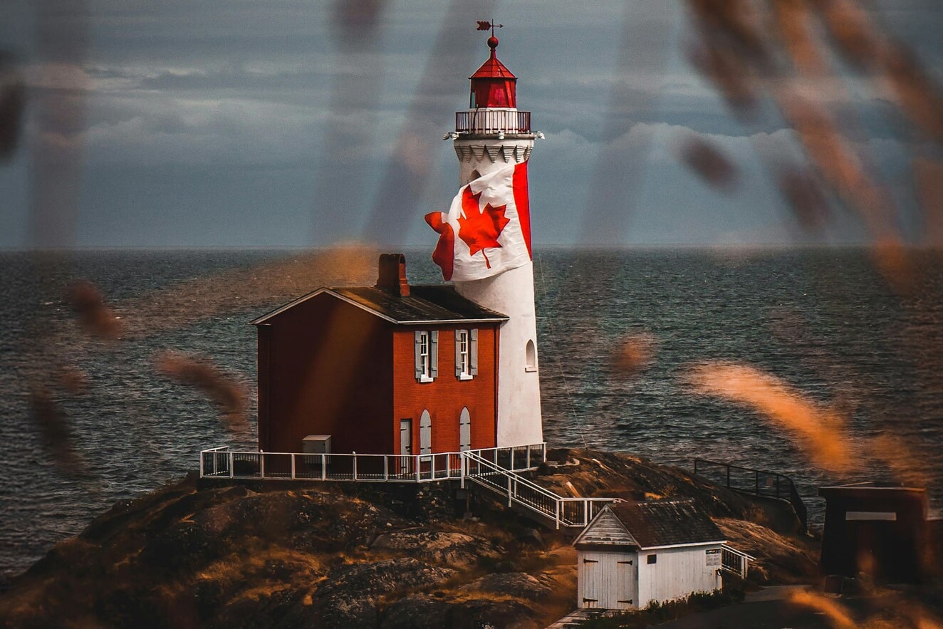 A lighthouse painted with a large Canadian flag stands on a rocky coast, perhaps prompting the question: Will 2025 be remembered as the year Canadians re-embraced nationalism? Red and white structures sit by the water under a cloudy sky and blurred grasses.
