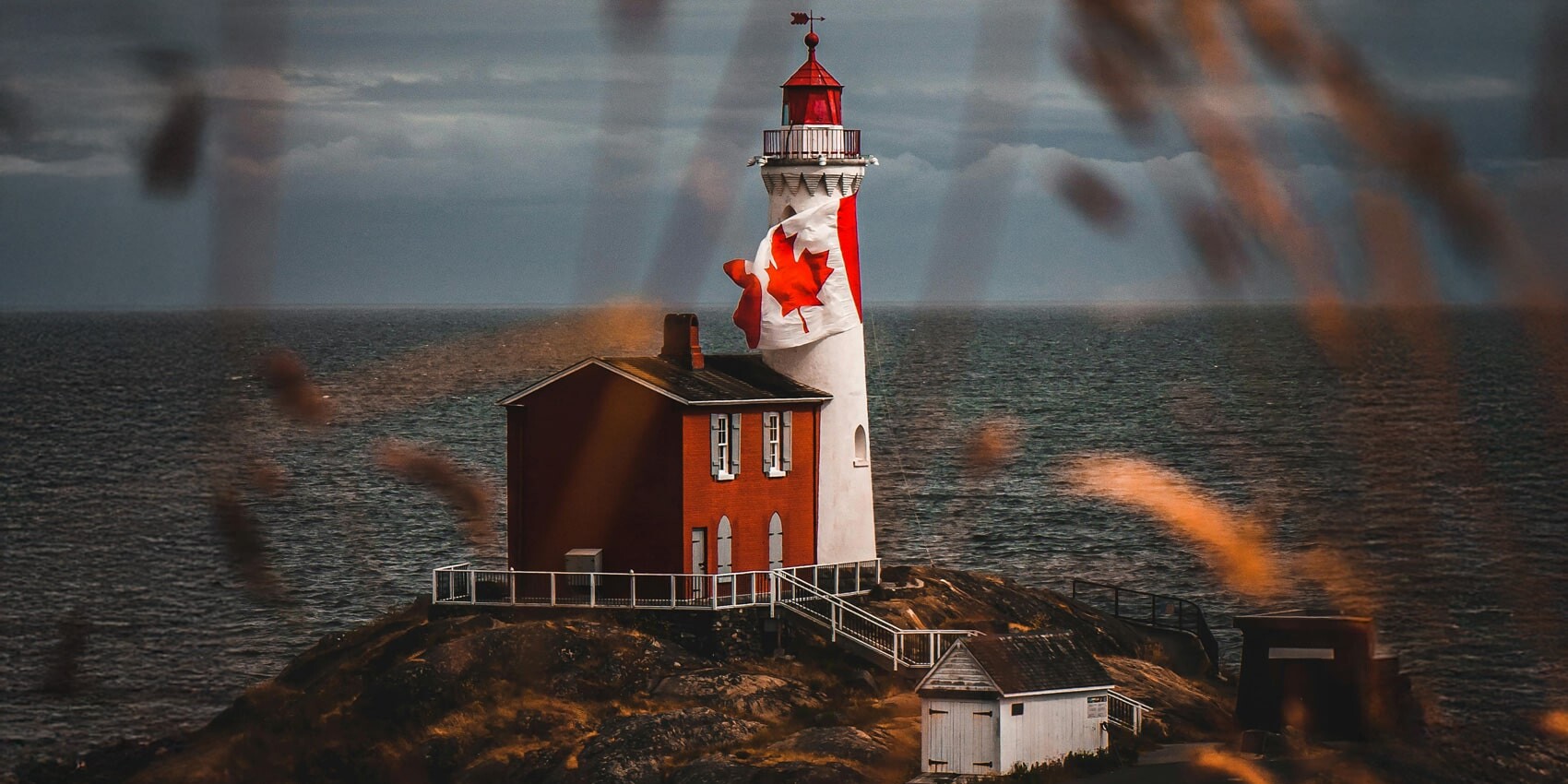 A lighthouse painted with a large Canadian flag stands on a rocky coast, perhaps prompting the question: Will 2025 be remembered as the year Canadians re-embraced nationalism? Red and white structures sit by the water under a cloudy sky and blurred grasses.
