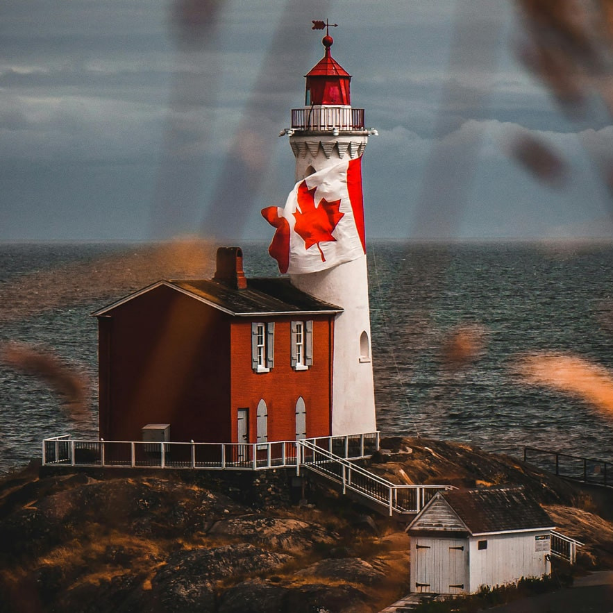 A lighthouse painted with a large Canadian flag stands on a rocky coast, perhaps prompting the question: Will 2025 be remembered as the year Canadians re-embraced nationalism? Red and white structures sit by the water under a cloudy sky and blurred grasses.
