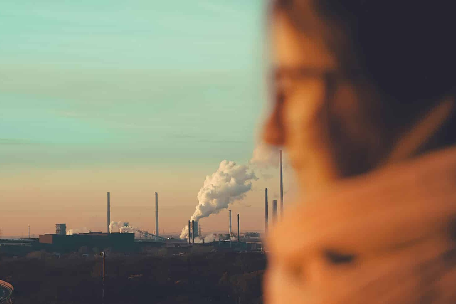 A blurred person in the foreground looks toward an industrial landscape with smokestacks, evoking the debate over climate action as most Canadians don't want a province-first approach to climate change under a pastel sunset sky.