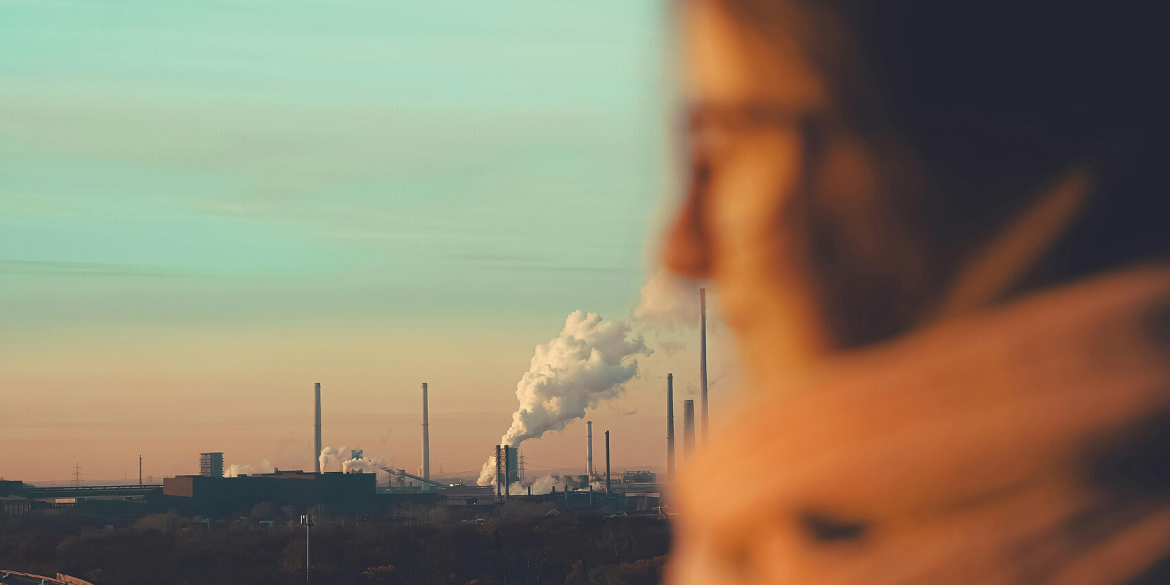 A blurred person in the foreground looks toward an industrial landscape with smokestacks, evoking the debate over climate action as most Canadians don't want a province-first approach to climate change under a pastel sunset sky.