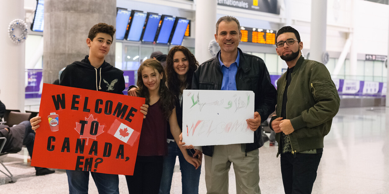 A group of five people at an airport hold welcome signs, including one that reads Welcome to Canada with Canadian flags. Smiling near arrival screens, they reflect how Canada must continue to model its refugee efforts on its Syrian crisis response.