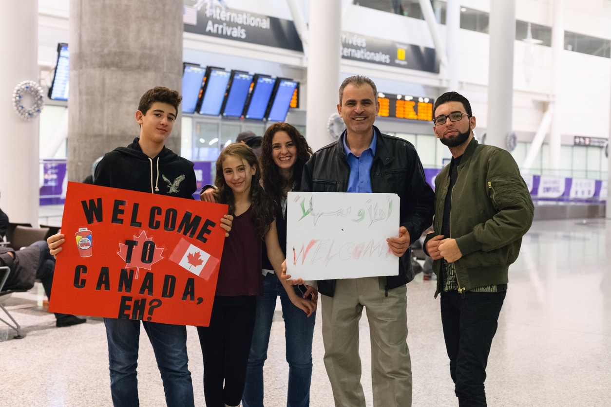 A group of five people at an airport hold welcome signs, including one that reads Welcome to Canada with Canadian flags. Smiling near arrival screens, they reflect how Canada must continue to model its refugee efforts on its Syrian crisis response.