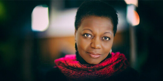 A woman with short hair, wearing a bright red scarf and dark earrings, looks softly at the camera. The blurred lights behind her subtly highlight the message: Yes Canada, we too have an anti-Black racism problem.