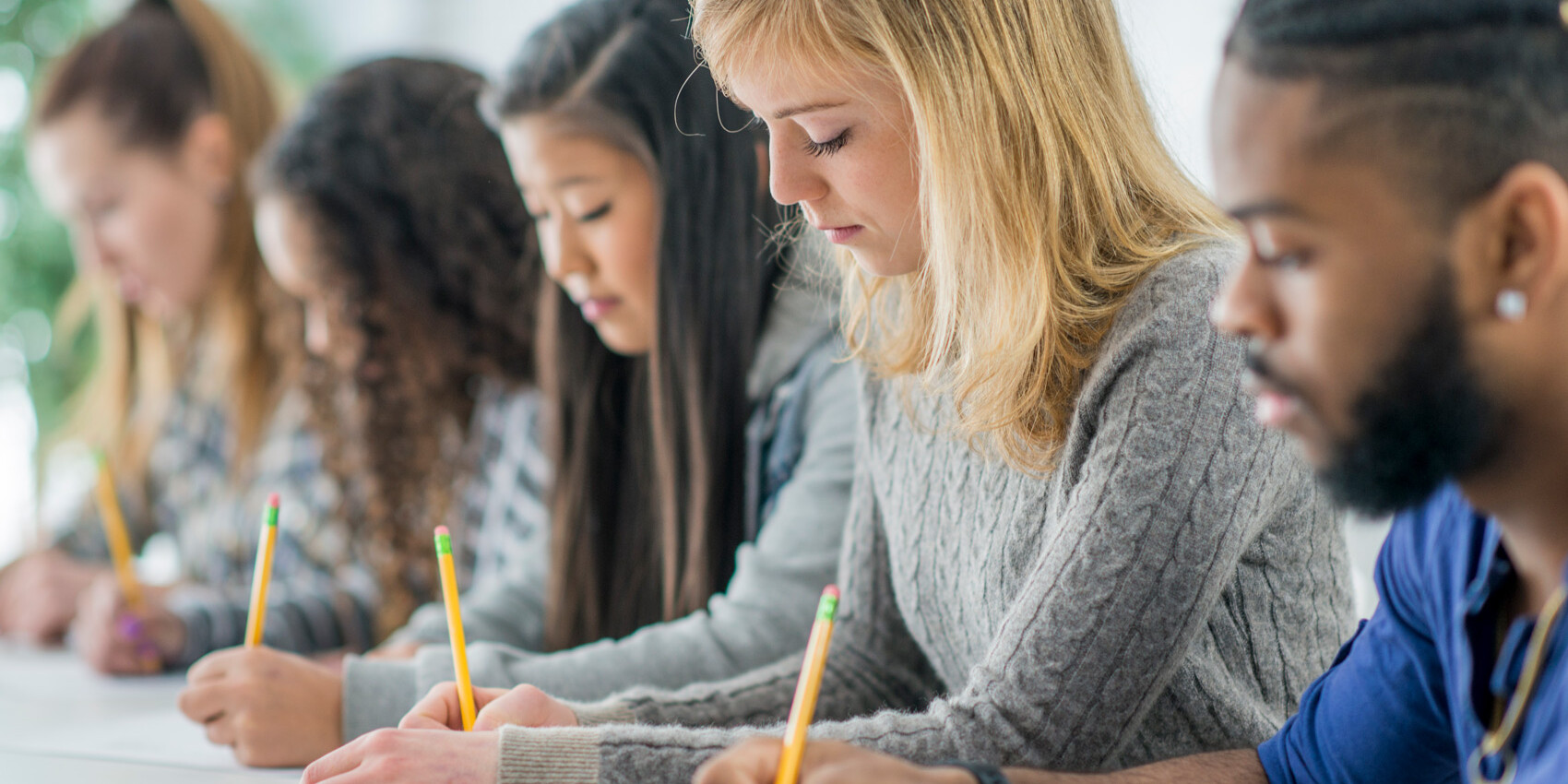 A diverse group of young adults sits in a row at a table, focused on writing with pencils on paper, reflecting how in Canada education excellence is also about equity within classroom settings.