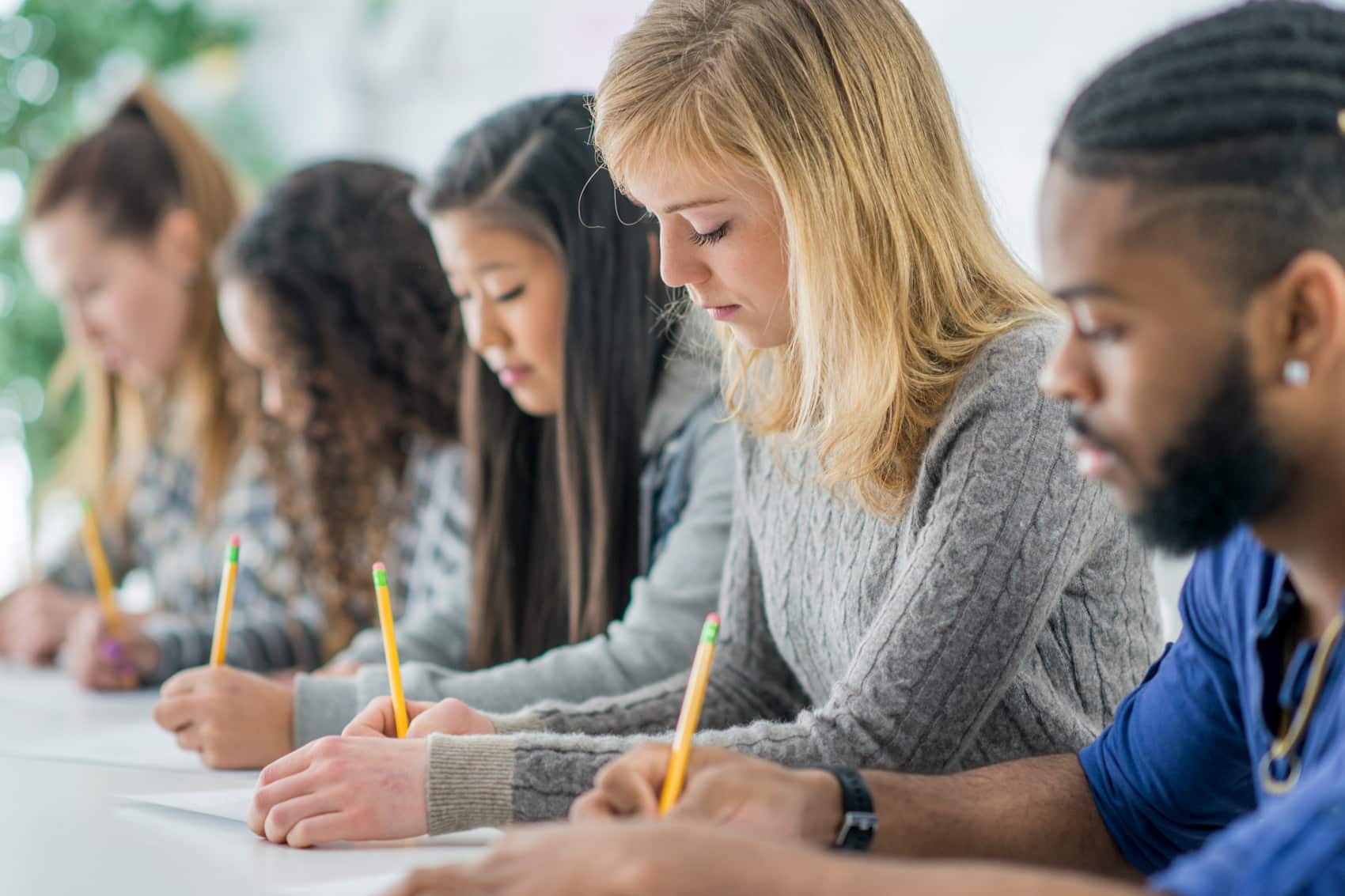 A diverse group of young adults sits in a row at a table, focused on writing with pencils on paper, reflecting how in Canada education excellence is also about equity within classroom settings.