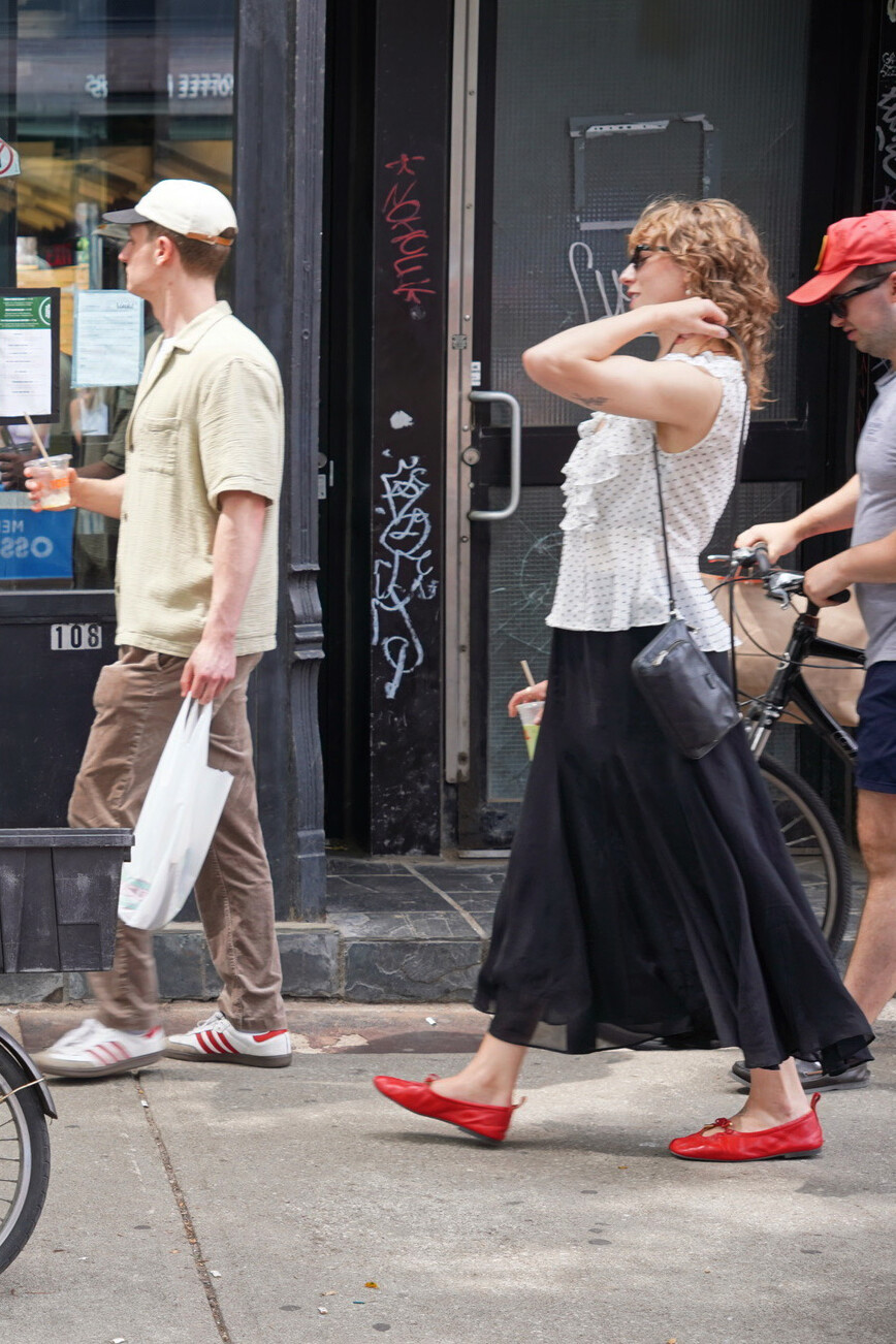 Three people walk on a city sidewalk, perhaps discussing what public opinion tells us about the political outlook for 2026. The woman in the center wears a white top, long black skirt, and red shoes, holding a drink. A bike is partially visible on the right.