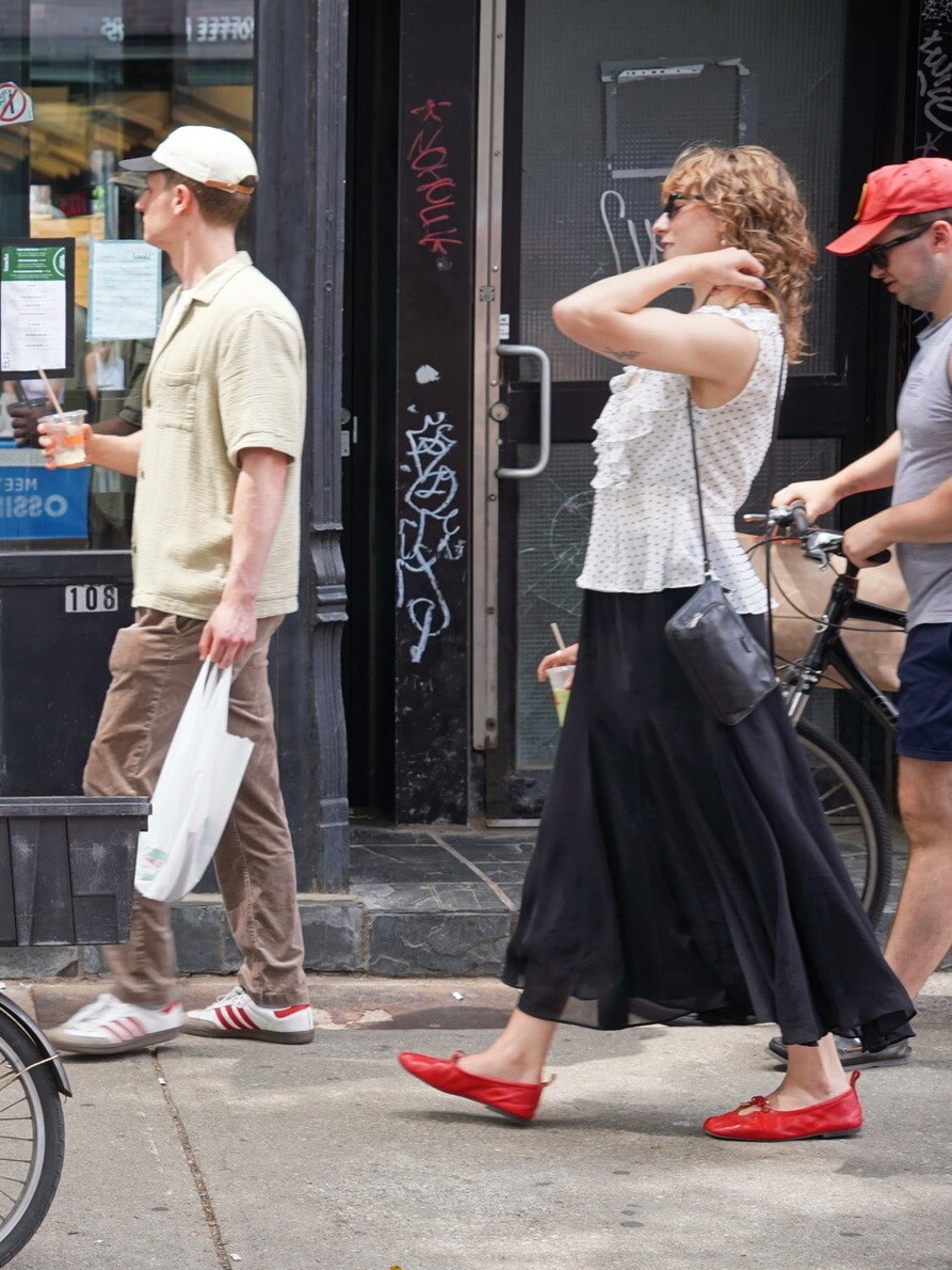 Three people walk on a city sidewalk, perhaps discussing what public opinion tells us about the political outlook for 2026. The woman in the center wears a white top, long black skirt, and red shoes, holding a drink. A bike is partially visible on the right.