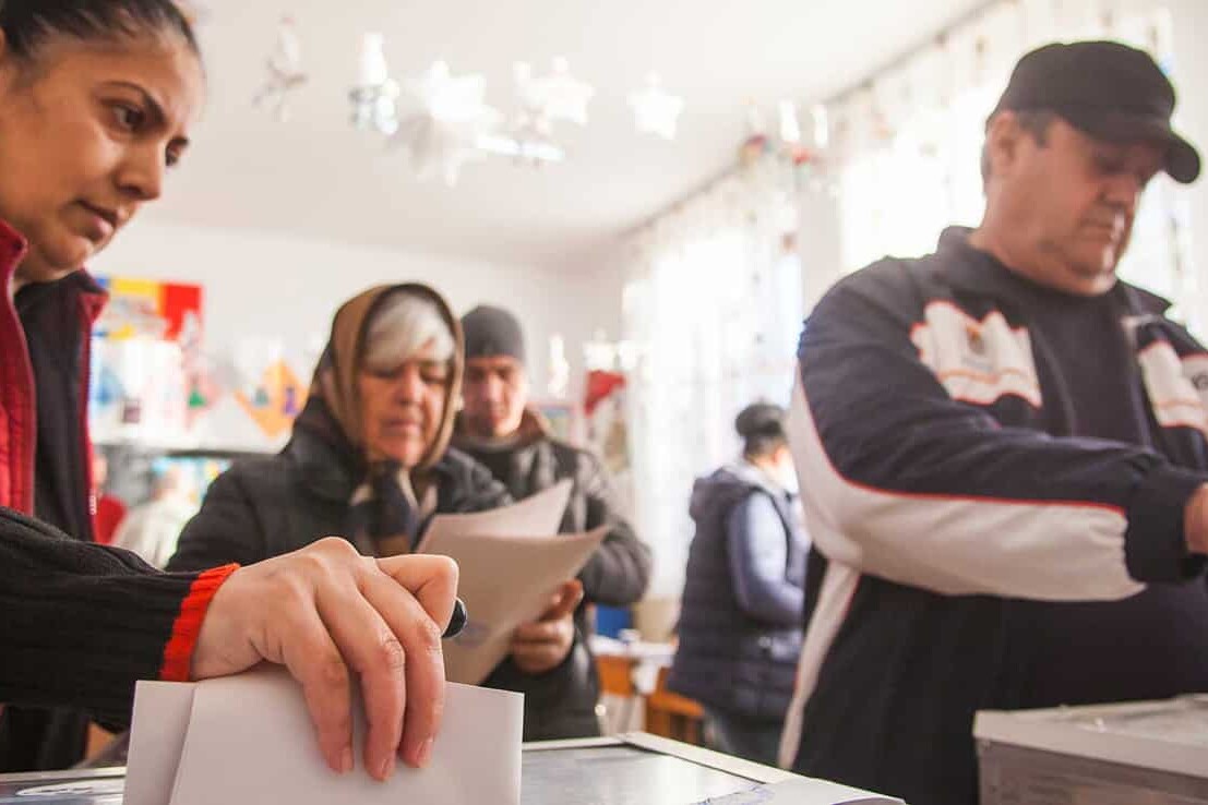 Several people stand in line indoors, casting ballots into clear boxes—an orderly scene that proves, despite angry protestors, Canadians still trust in our democracy. The focused atmosphere reflects faith in the election process.