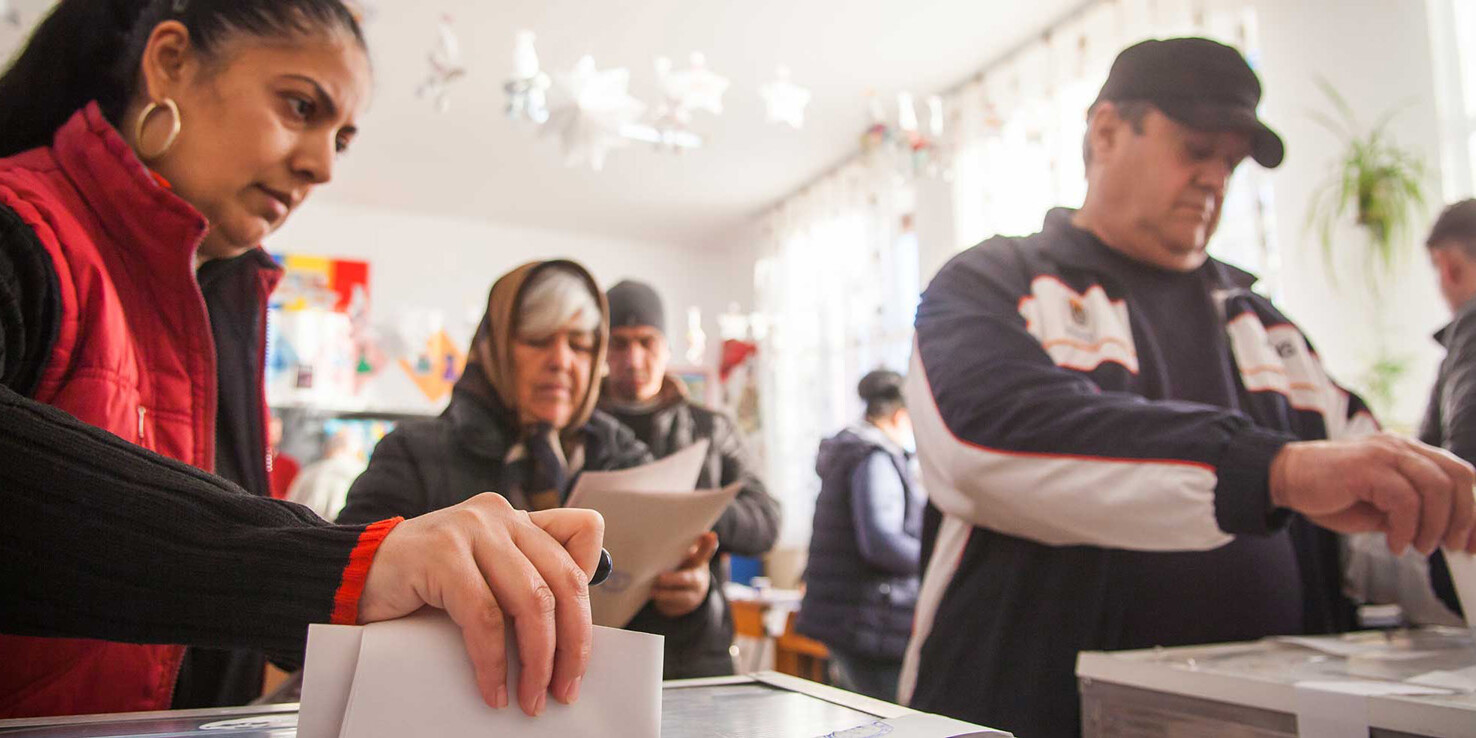 Several people stand in line indoors, casting ballots into clear boxes—an orderly scene that proves, despite angry protestors, Canadians still trust in our democracy. The focused atmosphere reflects faith in the election process.