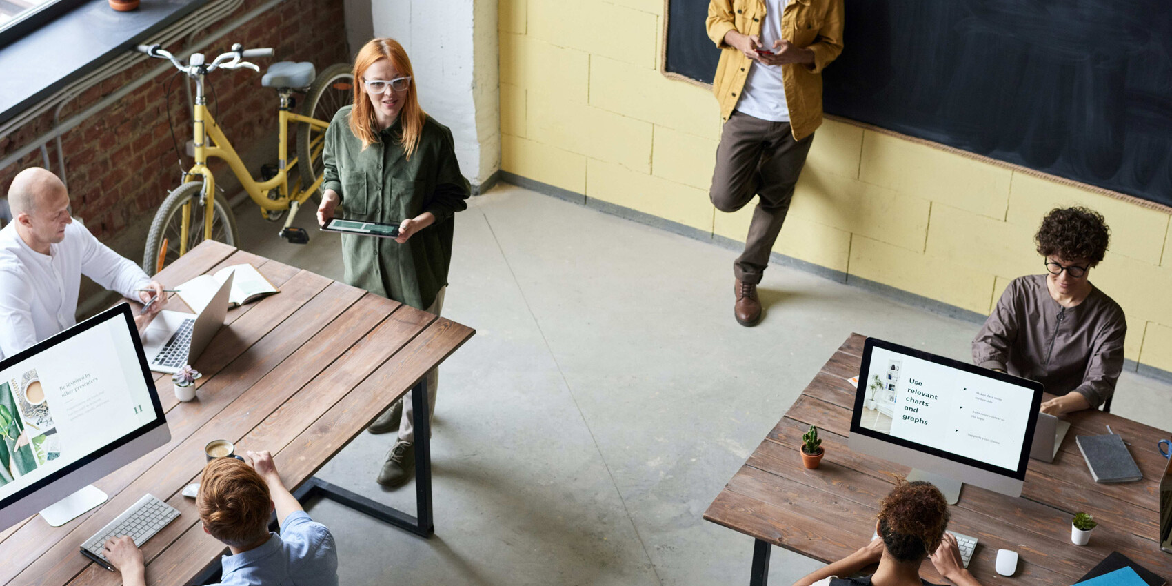 Five people work in a modern office with wooden desks and computers; one stands with a tablet, another leans against a wall on their phone discussing how Canada needs a bolder approach to skills training, while a yellow bicycle sits by large windows.
