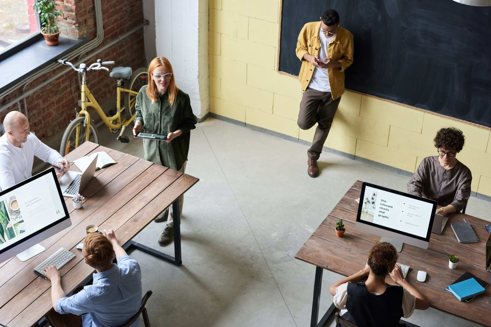 Five people work in a modern office with wooden desks and computers; one stands with a tablet, another leans against a wall on their phone discussing how Canada needs a bolder approach to skills training, while a yellow bicycle sits by large windows.